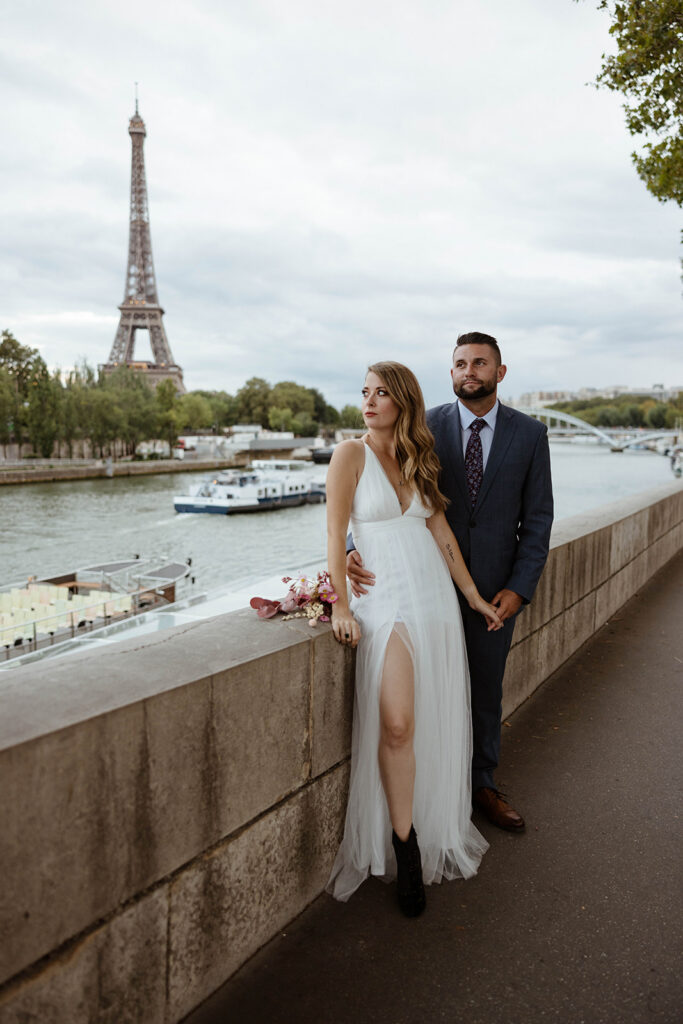 Ashley and Danny leaning on a stone wall along the Seine with the Eiffel Tower behind them on their Paris elopement day, photographed by Boston elopement photographer and officiant Erin of Boston